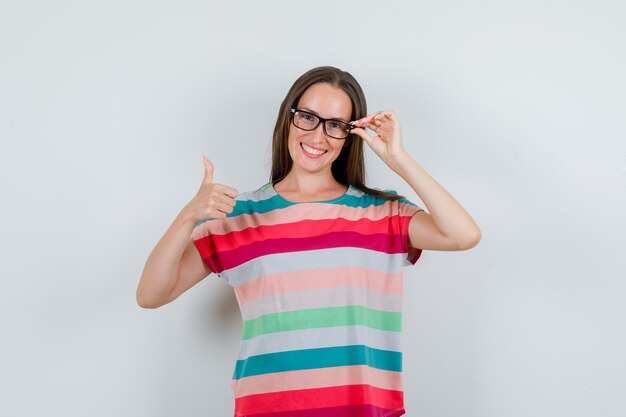 Jovem mulher aparecendo o polegar em uma camiseta, óculos e parecendo alegre. vista frontal.