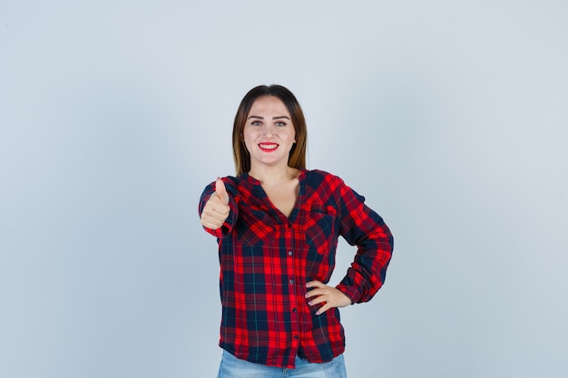 Jovem mulher aparecendo o polegar na camisa e olhando alegre, vista frontal.