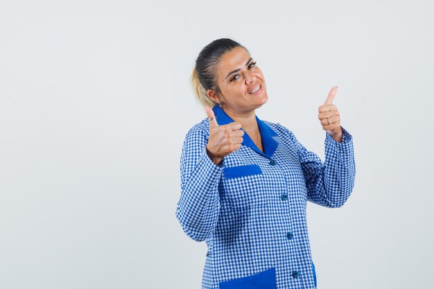 Jovem mulher aparecendo os polegares com ambas as mãos na camisa do pijama riscado azul e bonita vista frontal.