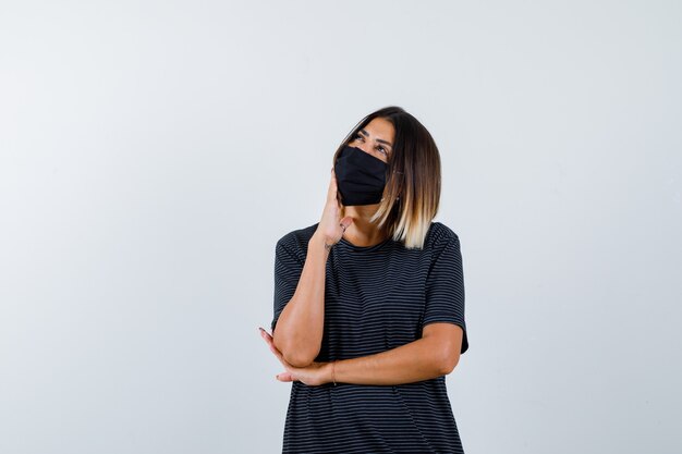 Jovem mulher apoiando o queixo na palma da mão, pensando em algo de vestido preto, máscara preta e olhando pensativa, vista frontal.