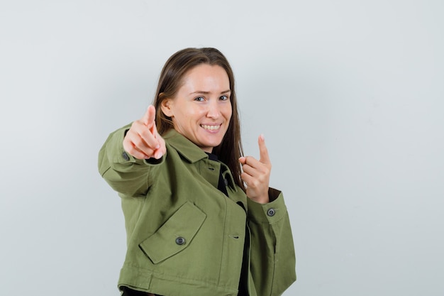 Jovem mulher apontando com jaqueta verde e olhando alegre, vista frontal.