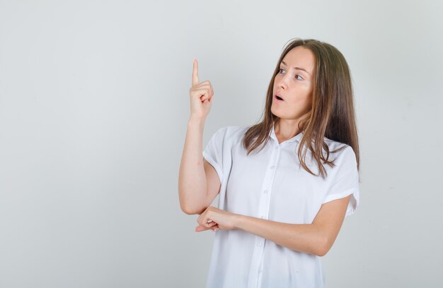 Jovem mulher apontando o dedo para cima em uma camisa branca, parecendo confusa