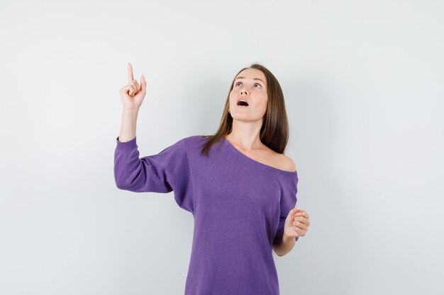 Jovem mulher apontando o dedo para cima na camisa violeta e olhando com foco, vista frontal.