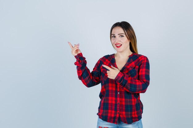 Jovem mulher apontando para a esquerda com os dedos indicadores em camisa quadrada, jeans e olhando alegre, vista frontal.