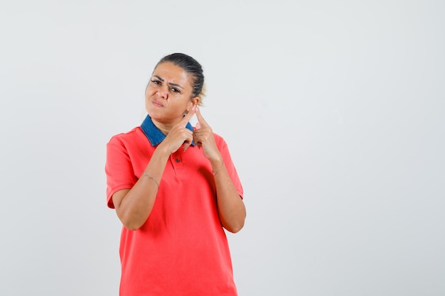 Jovem mulher apontando para a orelha em t-shirt vermelha e parecendo irritada, vista frontal.