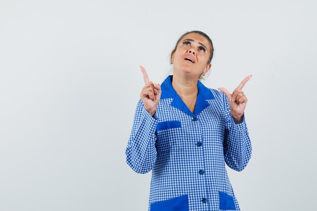 Jovem mulher apontando para cima com o dedo indicador em uma camisa de pijama azul guingão e olhando pensativa, vista frontal.