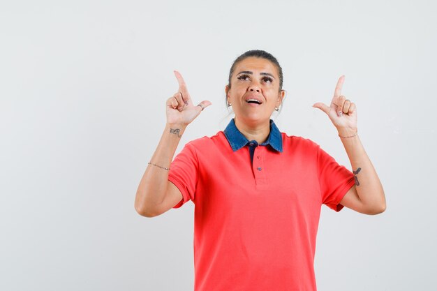 Jovem mulher apontando para cima com os dedos indicadores em t-shirt vermelha e olhando surpresa, vista frontal.