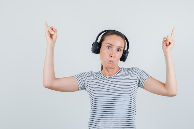 Jovem mulher apontando para cima em t-shirt, fones de ouvido e parecendo perplexa. vista frontal.