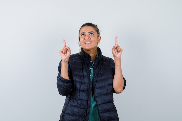 Jovem mulher apontando para cima, olhando para cima com uma jaqueta inflável e parecendo alegre. vista frontal.