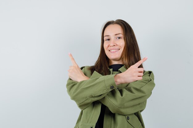 Jovem mulher apontando para o lado com os braços cruzados numa jaqueta verde e parecendo confusa. vista frontal.