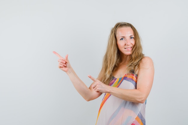 Jovem mulher apontando para o lado com os dedos para cima com vestido de verão e olhando feliz.