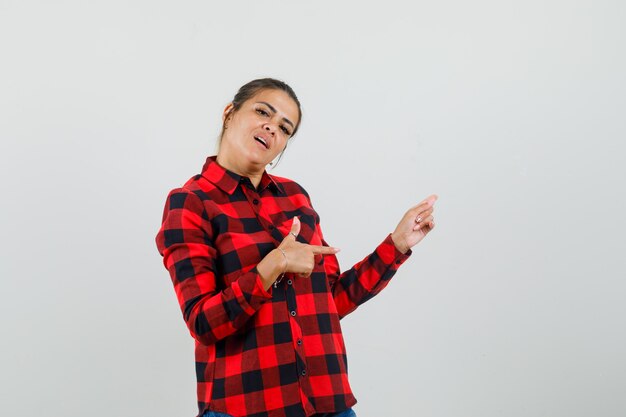 Jovem mulher apontando para o lado em camisa xadrez e olhando confiante, vista frontal.