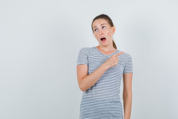 Jovem mulher apontando para o lado em t-shirt e parecendo confusa. vista frontal.