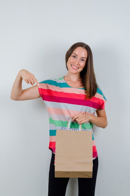 Jovem mulher apontando para sacos de papel em t-shirt, calças e olhando feliz, vista frontal.