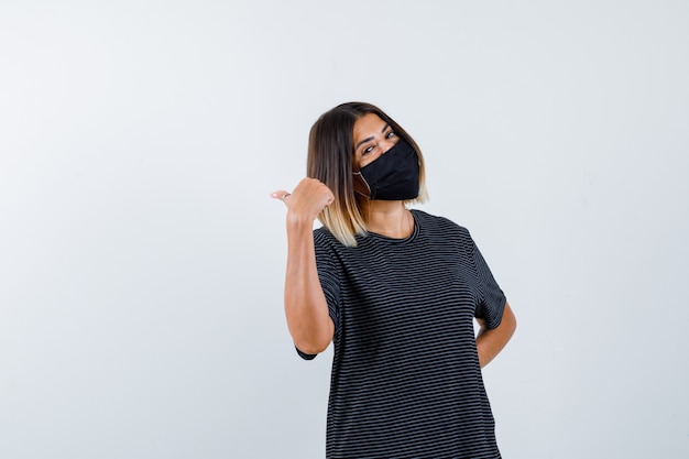 Jovem mulher apontando para trás, segurando a mão atrás da cintura em um vestido preto, máscara preta e parecendo feliz. vista frontal.