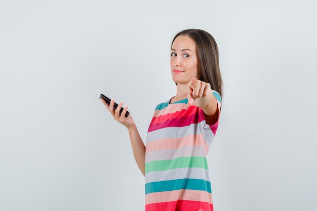 Jovem mulher apontando para você enquanto segura o smartphone na vista frontal de t-shirt.