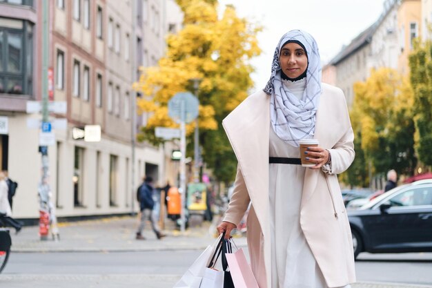 Jovem mulher árabe atraente em hijab andando pela rua da cidade com sacolas de compras e café