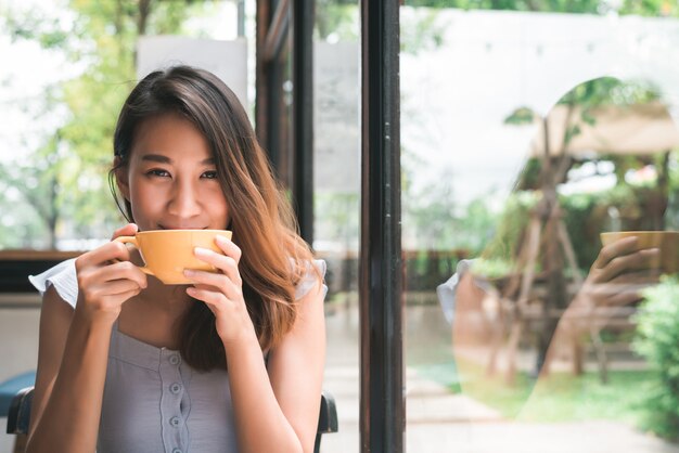 Jovem mulher asiática alegre que bebe o café ou o chá morno que aprecia a ao sentar-se no café