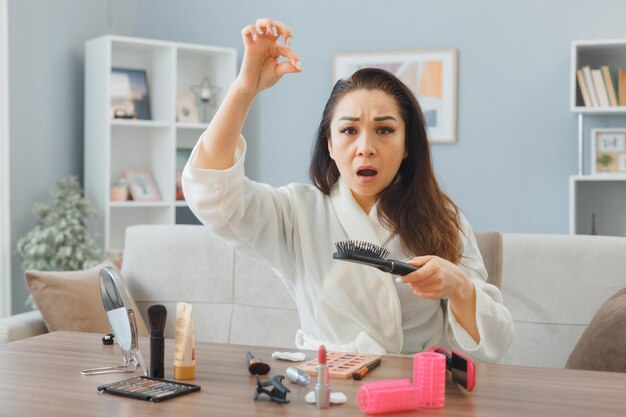 Jovem mulher asiática com toalha de roupão de banho sentado na penteadeira em casa interior escovando o cabelo sendo confuso e chateado com perda de cabelo fazendo rotina de maquiagem matinal