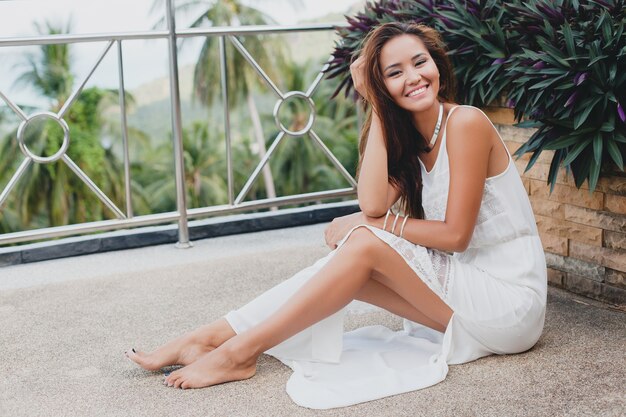 Jovem mulher asiática elegante sentada no chão com um vestido boho branco, estilo vintage, natural, sorridente, feliz, magra, férias tropicais, hotel