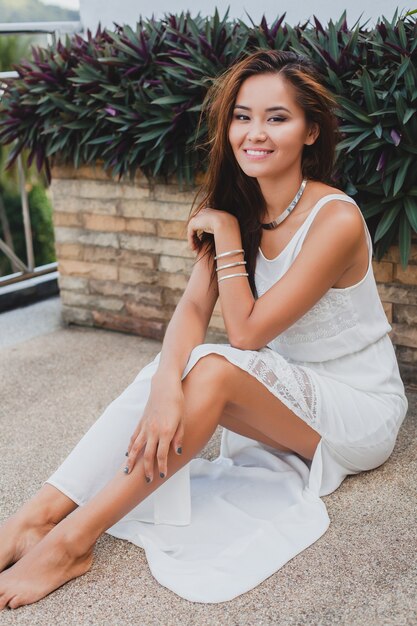 Jovem mulher asiática elegante sentada no chão com um vestido boho branco, estilo vintage, natural, sorridente, feliz, magra, férias tropicais, hotel