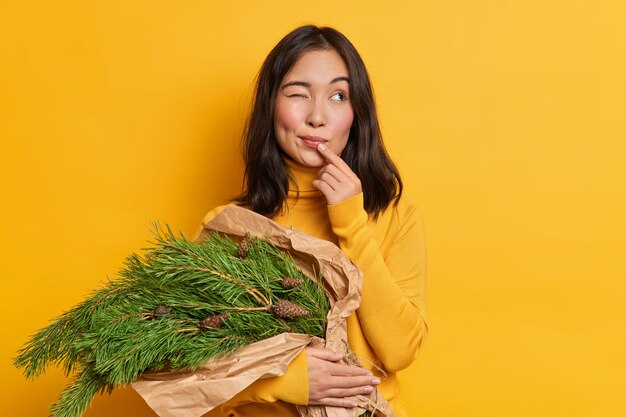 Jovem mulher asiática pensativa, com um olho fechado, carregando um buquê de ramos com pinhas, pensa em como celebrar poses de ano novo dentro de casa