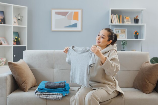 Jovem mulher asiática sentada em um sofá no interior de casa segurando a pilha de roupa limpa feliz e positiva sorrindo