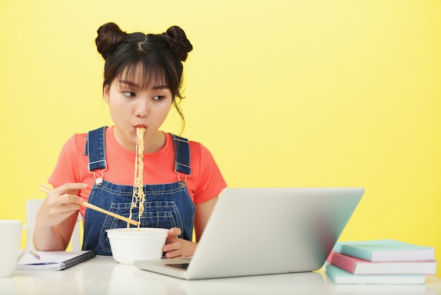 Jovem mulher asiática sentada na mesa, comendo macarrão com pauzinhos e olhando para laptop