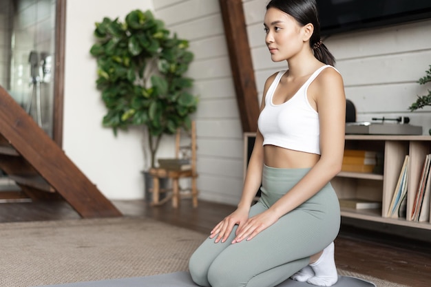 Jovem mulher asiática treino em casa meditando ou fazendo exercícios de ioga na sala de estar usando esportes w.