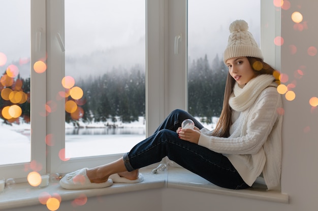 Jovem mulher atraente em um elegante suéter branco de malha, lenço e chapéu sentada em casa no parapeito da janela no Natal segurando a decoração do presente da bola de neve de vidro, vista da floresta de inverno, luzes bokeh