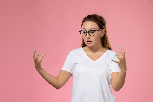 Jovem mulher atraente em uma camiseta branca posando de frente para mostrar algo com as mãos ao som de música no fundo rosa