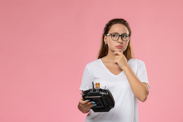 Jovem mulher atraente em uma camiseta branca segurando o controle remoto com expressão pensativa no fundo rosa.