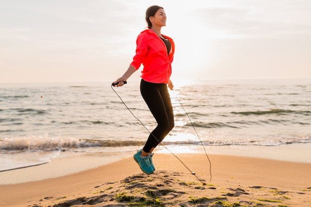 Jovem mulher atraente fazendo exercícios esportivos ao nascer do sol da manhã na praia do mar em roupas esportivas, estilo de vida saudável, ouvindo música em fones de ouvido, vestindo uma jaqueta corta-vento rosa, pulando corda