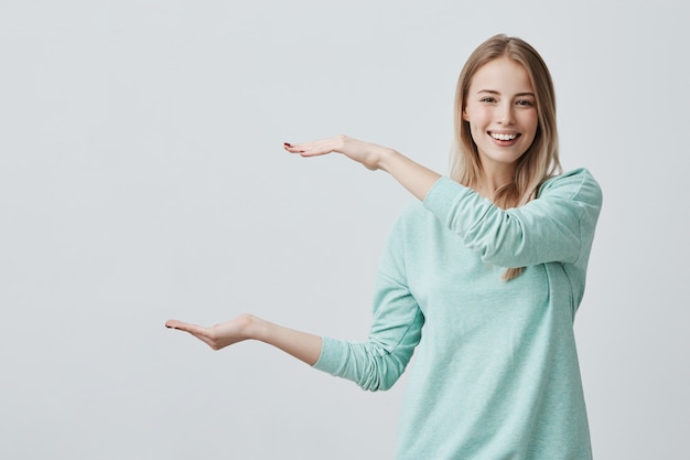 Jovem mulher atraente feliz na camisola azul clara, mostrando algo grande com as mãos