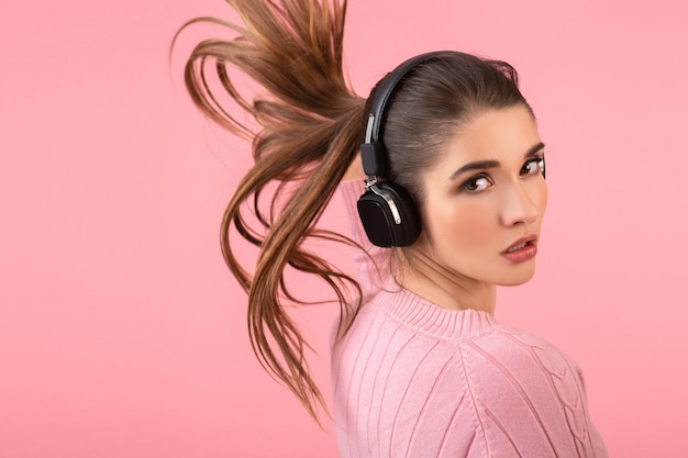 Jovem mulher atraente ouvindo música em fones de ouvido sem fio, vestindo um suéter rosa, sorrindo, feliz humor positivo, posando em fundo rosa