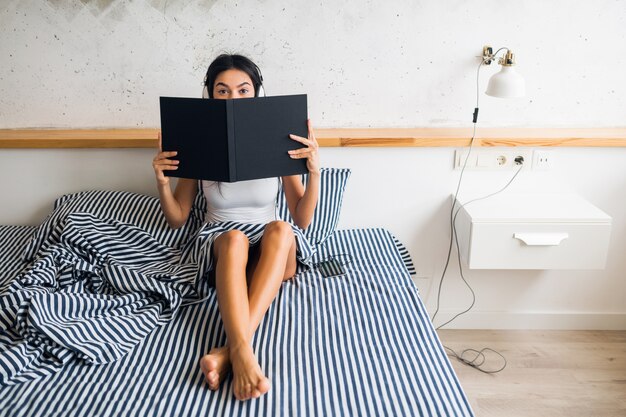 Jovem mulher atraente sentada na cama de pijama, sorrindo, quarto, emoção feliz, acordar de manhã, ouvindo música em fones de ouvido, lendo um livro, segurando um álbum, escondendo-se, expressão de surpresa