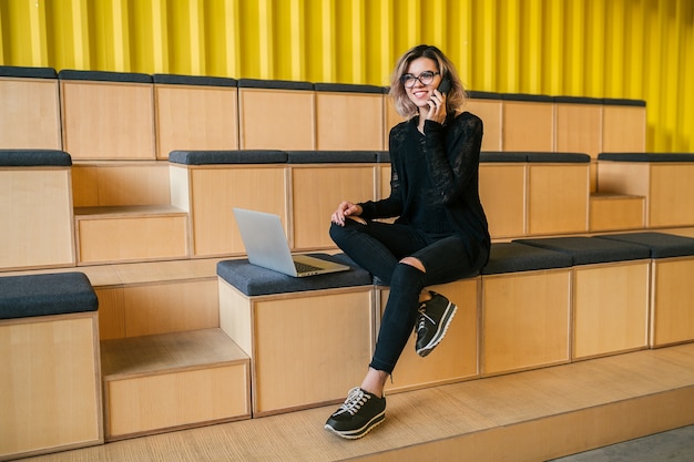 Jovem mulher atraente sentada na sala de aula, trabalhando no laptop, usando óculos, auditório moderno, educação do aluno online, freelancer, sorrindo, falando ao telefone