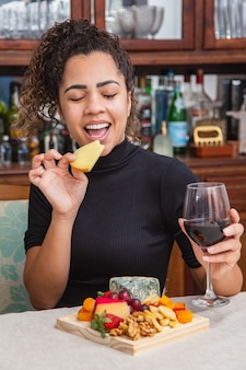 Jovem mulher bebendo vinho e comendo queijo sozinha na sala de estar. mulher comendo salgadinhos de queijo e bebendo vinho para relaxar depois do trabalho.