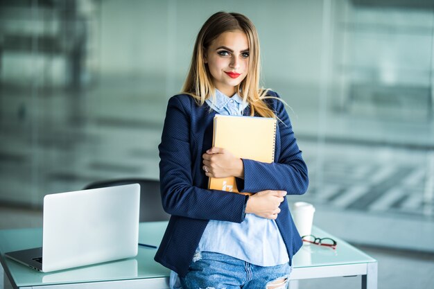 Jovem mulher bem sucedida em roupas casuais, segurando o trabalho do caderno em pé perto da mesa branca com o laptop no escritório. Conceito de carreira empresarial de realização.