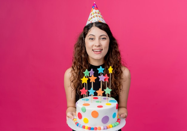 Jovem mulher bonita com cabelo encaracolado com um boné de férias segurando um bolo de aniversário feliz e animada sorrindo conceito de festa de aniversário em pé sobre a parede rosa