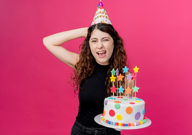 Jovem mulher bonita com cabelo encaracolado com um boné de férias segurando um bolo de aniversário, sorrindo alegremente, parecendo feliz e alegre conceito de festa de aniversário em pé sobre a parede rosa