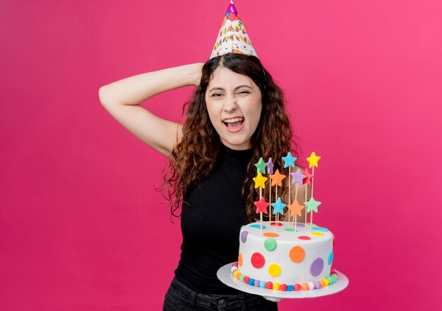 Jovem mulher bonita com cabelo encaracolado com um boné de férias segurando um bolo de aniversário, sorrindo alegremente, parecendo feliz e alegre conceito de festa de aniversário em pé sobre a parede rosa