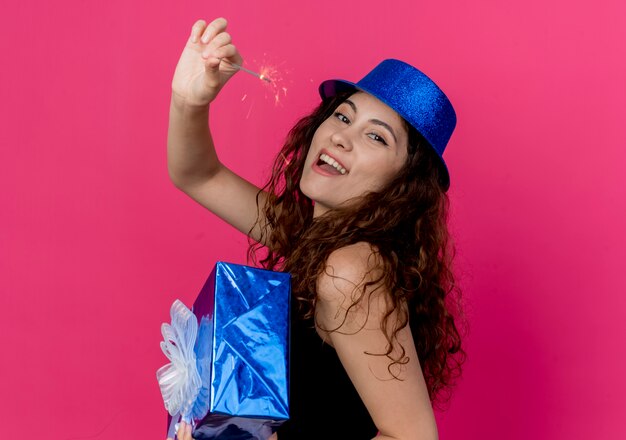 Jovem mulher bonita com cabelo encaracolado com um chapéu de férias segurando uma caixa de presente de aniversário e um diamante feliz e animado conceito de festa de aniversário em pé sobre a parede rosa