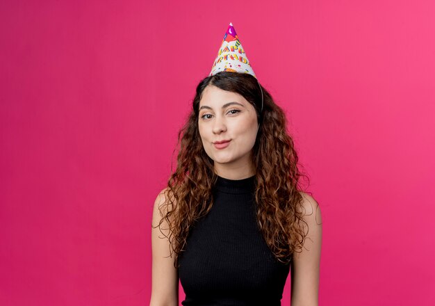 Jovem mulher bonita com cabelo encaracolado em um boné de férias feliz e positiva sorrindo conceito de festa de aniversário em pé sobre a parede rosa