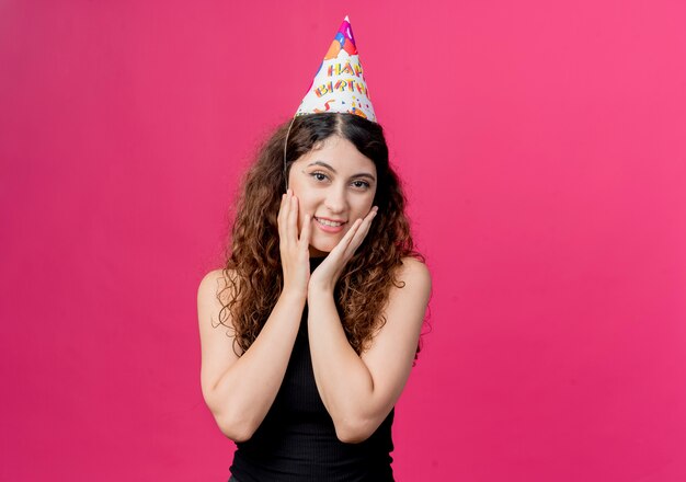 Jovem mulher bonita com cabelo encaracolado em um boné de férias feliz e surpresa segurando o rosto com braços conceito de festa de aniversário em pé sobre a parede rosa