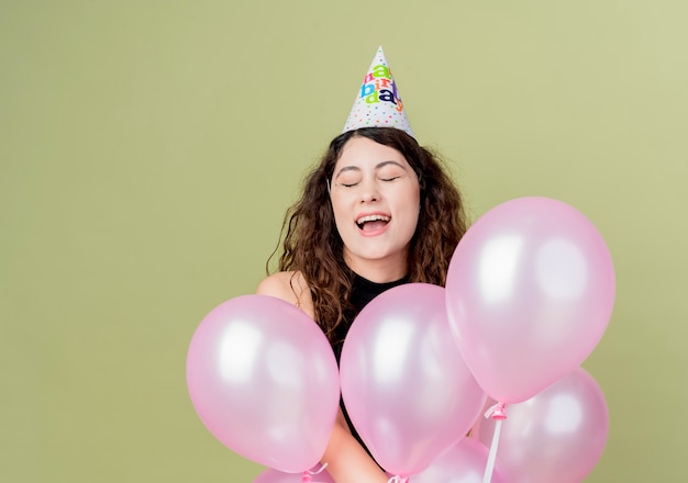 Jovem mulher bonita com cabelo encaracolado em um boné de férias segurando balões de ar feliz e animada conceito de festa de aniversário em pé sobre a parede de luz