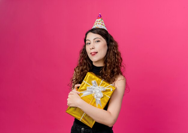 Jovem mulher bonita com cabelo encaracolado em um boné de férias segurando uma caixa de presente de aniversário sorrindo alegremente esticando a língua para fora conceito de festa de aniversário em pé sobre a parede rosa
