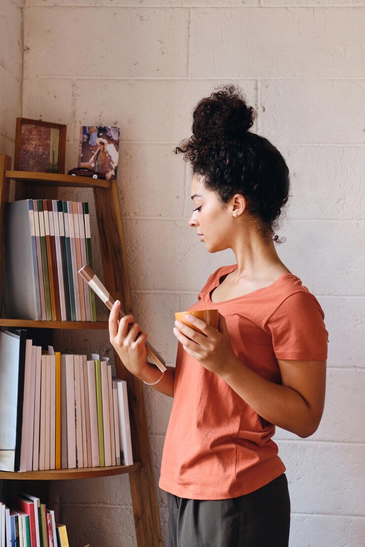Foto grátis jovem mulher bonita com cabelo encaracolado escuro na camiseta segurando a xícara de café na mão e escolhendo cuidadosamente o livro da estante na casa moderna e aconchegante