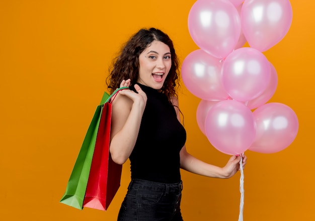 Foto grátis jovem mulher bonita com cabelo encaracolado segurando um monte de balões de ar e sacos de papel feliz e alegre sorrindo conceito de festa de aniversário em pé sobre a parede laranja