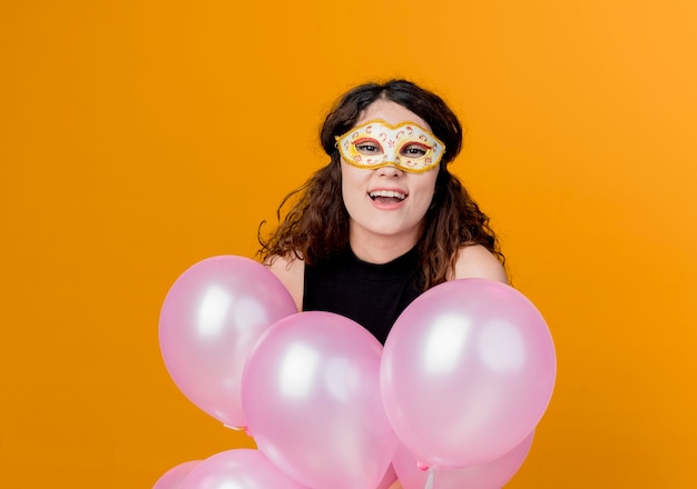 Jovem mulher bonita com cabelo encaracolado segurando um monte de balões de ar na máscara de festa conceito de festa de aniversário feliz e alegre sobre laranja